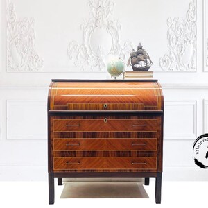 May include: A vintage wooden roll top desk with three drawers and a dark wood finish. The desk has a decorative inlay of light wood and brass accents. The desk is in front of a white wall with decorative molding.