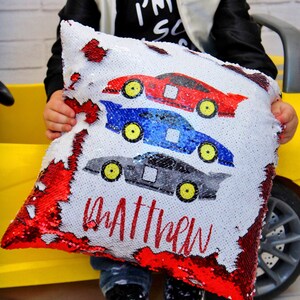 May include: A red, white, and blue sequin pillow with a design of three race cars stacked on top of each other. The pillow has the name "Matthew" written on it in red.