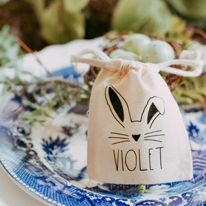 May include: A white fabric drawstring bag with a bunny rabbit design and the name "Violet" printed on it. The bag is sitting on a blue and white patterned plate.