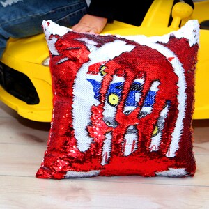 May include: Red and white sequin pillow with a blue and white graphic of a robot. The pillow is sitting on a yellow toy car.