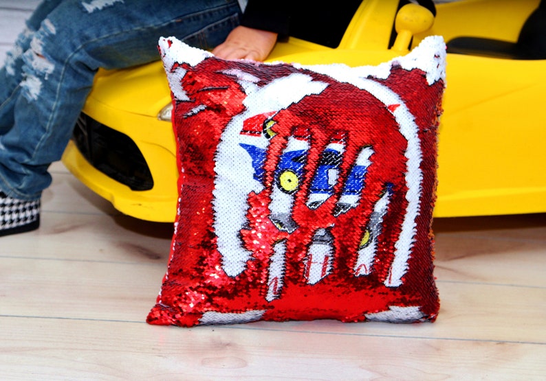 May include: Red and white sequin pillow with a blue and white graphic of a robot. The pillow is sitting on a yellow toy car.