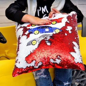May include: A red, white, and blue sequin pillow with a reversible design. One side features three cartoon cars, and the other side is solid red. The pillow is being held by a child.