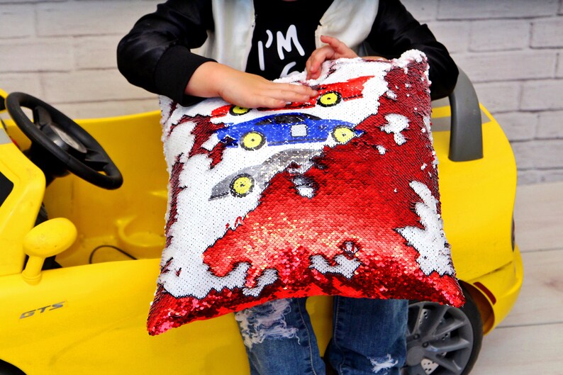 May include: A red, white, and blue sequin pillow with a reversible design. One side features three cartoon cars, and the other side is solid red. The pillow is being held by a child.