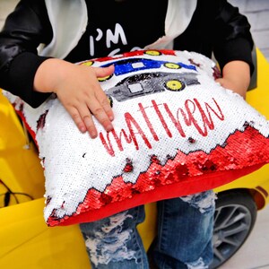May include: A white and red sequin pillow with a red car design and the name "Matthew" written in red. The pillow is being held by a child.