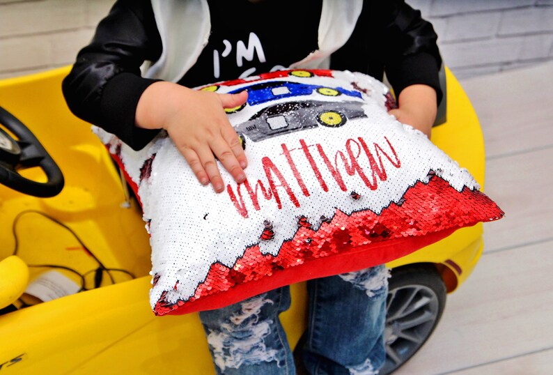 May include: A white and red sequin pillow with a red car design and the name "Matthew" written in red. The pillow is being held by a child.