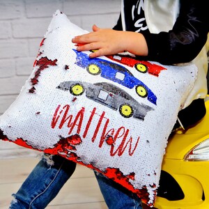 May include: A white pillow with red, blue, and gray race cars and the name "Matthew" in red lettering. The pillow is made of sequins that can be flipped to reveal a different color.