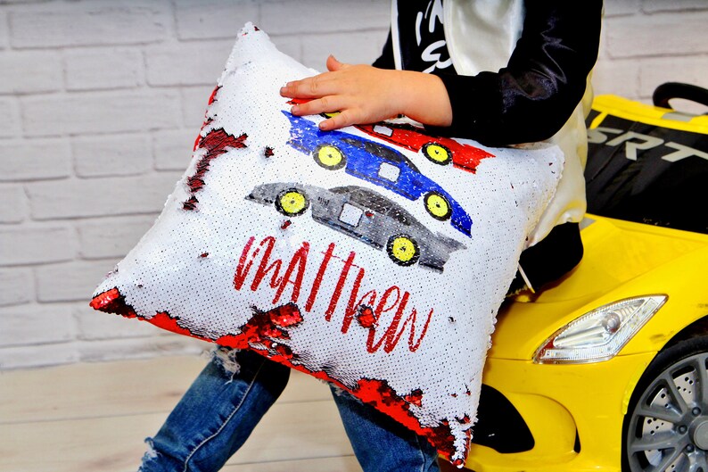 May include: A white pillow with red, blue, and gray race cars and the name "Matthew" in red lettering. The pillow is made of sequins that can be flipped to reveal a different color.