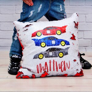 May include: A white pillow with a red, blue, and gray race car design. The pillow is made of sequins and has the name "Matthew" written on it.