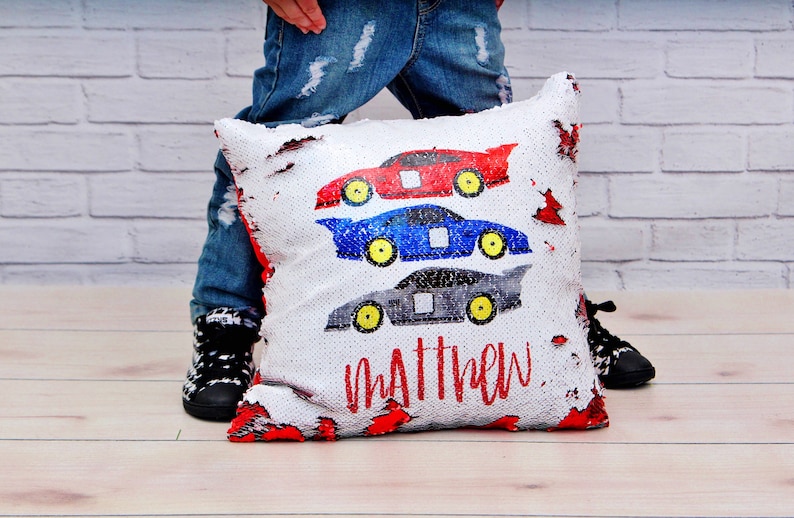 May include: A white pillow with a red, blue, and gray race car design. The pillow is made of sequins and has the name "Matthew" written on it.