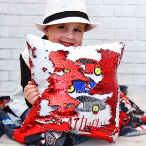 May include: A child wearing a white hat holds a red and white sequin pillow with a colorful car design. The pillow has the words "Little Dude" printed on it.