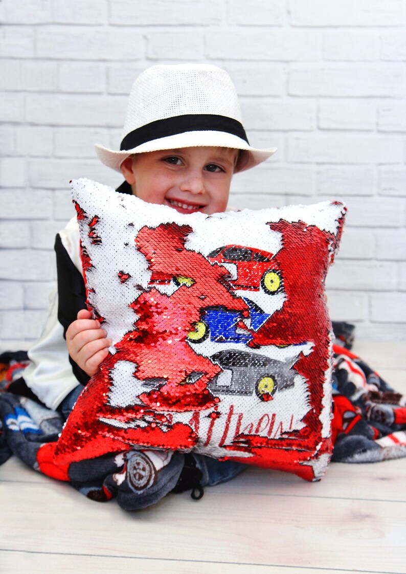 May include: A child wearing a white hat holds a red and white sequin pillow with a colorful car design. The pillow has the words "Little Dude" printed on it.