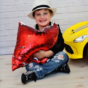 May include: A young person wearing a white fedora, black jacket, and ripped jeans is sitting on a wooden floor holding a red sequin pillow. A yellow toy car is behind the person.