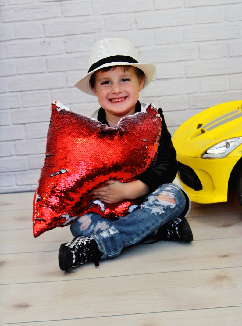 May include: A young person wearing a white fedora, black jacket, and ripped jeans is sitting on a wooden floor holding a red sequin pillow. A yellow toy car is behind the person.