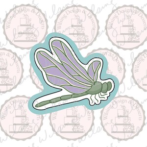 May include: A dragonfly-shaped cookie cutter with a light blue outline. The dragonfly has purple wings with green veins, a green head, and a segmented green body. The background features a repeating pattern of pink circular logos with text.