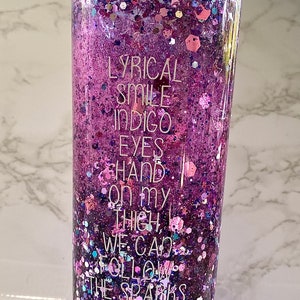 May include: A clear tumbler with a teal lid filled with purple, pink and blue glitter. The tumbler has the text "LYRICAL SMILE INDIGO EYES HAND ON MY THIGH WE CAN FOLLOW THE SPARKS" printed on the side.