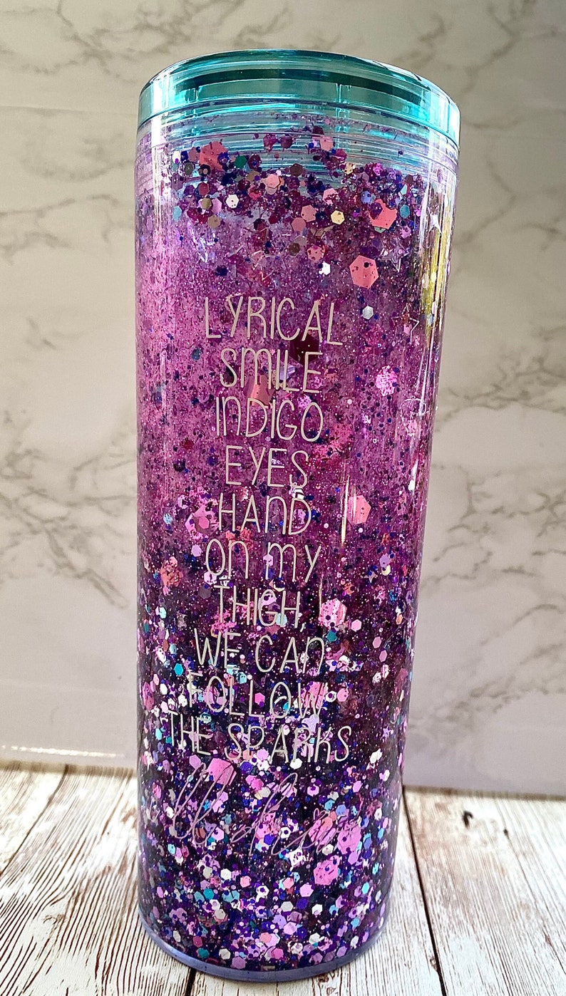 May include: A clear tumbler with a teal lid filled with purple, pink and blue glitter. The tumbler has the text "LYRICAL SMILE INDIGO EYES HAND ON MY THIGH WE CAN FOLLOW THE SPARKS" printed on the side.