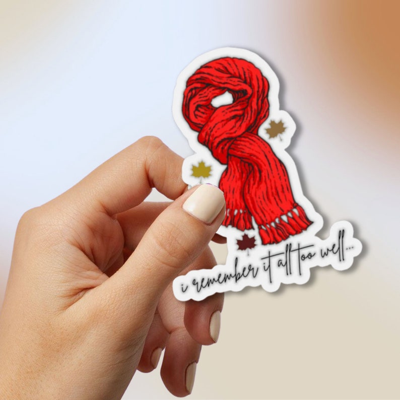 May include: A die-cut sticker featuring a bright red knitted scarf with fringe at the bottom, accented by small gold and dark brown maple leaf illustrations. The text "i remember it all too well..." is printed in a black cursive script at the base.