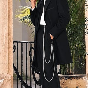 May include: A black suit jacket, white dress shirt, black tie, and black trousers paired with a silver pocket watch chain. The outfit includes a black fedora hat with a white band and a feather, and black and white wingtip dress shoes.