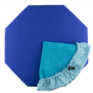 Puede incluir: Un cojín azul en forma de octógono con un ribete de tela vichy turquesa y blanco. El cojín tiene un borde con volantes y un pequeño botón negro.