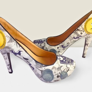 Puede incluir: Un par de zapatos de tac&oacute;n alto blancos con ribete negro. Los zapatos est&aacute;n cubiertos con un estampado de personajes de la pel&iacute;cula "Pesadilla antes de Navidad".