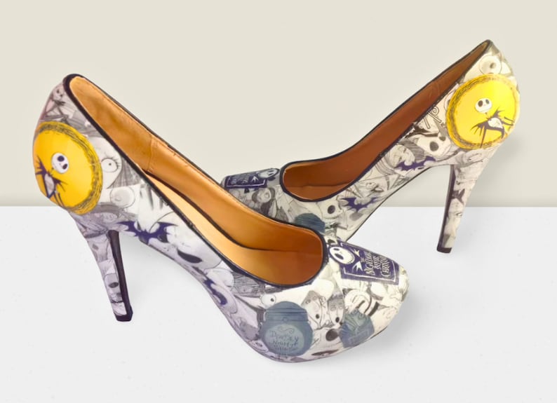 Puede incluir: Un par de zapatos de tac&oacute;n alto blancos con ribete negro. Los zapatos est&aacute;n cubiertos con un estampado de personajes de la pel&iacute;cula "Pesadilla antes de Navidad".