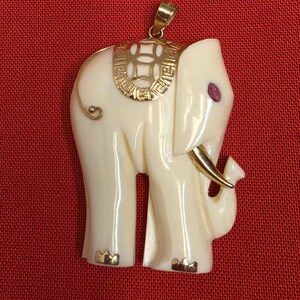 Puede incluir: Colgante de elefante de color marfil con detalles dorados. El colgante presenta una montura, colmillos y patas doradas. Una pequeña gema roja está engastada en el ojo del elefante. El colgante está suspendido de una anilla dorada. El fondo es rojo liso.