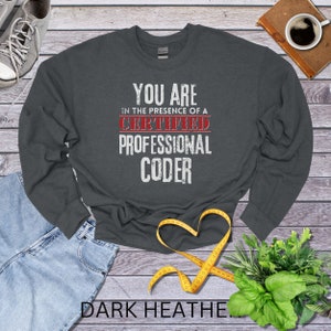 May include: Dark heather grey long-sleeved sweatshirt with the text "You are in the presence of a Certified Professional Coder" in white with red accents.