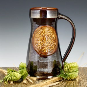 May include: A brown ceramic beer stein featuring a Celtic knot design. The stein has a wide base and a handle. It is resting on a wooden surface with wheat stalks and green hops.