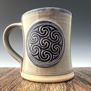 Puede incluir: Una taza de cerámica gris con un diseño de nudo celta azul y blanco. La taza tiene un asa redondeada y una base ancha.