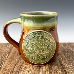 May include: A brown and green ceramic mug with a Celtic tree of life design. The mug has a rounded body and a handle.