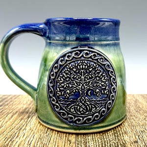 Puede incluir: Una taza de cerámica verde y azul con un diseño de árbol de la vida. La taza tiene un asa y un diseño en relieve de un árbol con raíces y ramas.