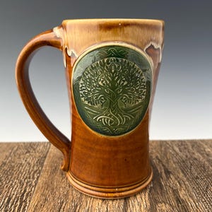 May include: A brown ceramic mug with a green tree of life design. The mug has a handle and is glazed in a brown color.