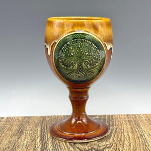 Puede incluir: Una copa de cerámica marrón con un diseño de árbol de la vida verde. La copa tiene un borde ancho con un esmalte marrón claro, un tallo detallado y una base redonda. El diseño del árbol está centrado en la copa.
