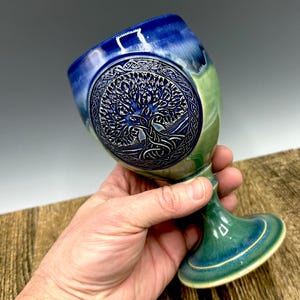 May include: A ceramic goblet with a Celtic tree of life design. The goblet features a blue and green color scheme, with a detailed tree carving on the bowl. The stem and base are a darker shade of green, creating a striking contrast.