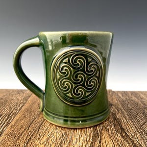 May include: A green ceramic mug with a Celtic knot design. The mug has a rounded handle and a wide base.