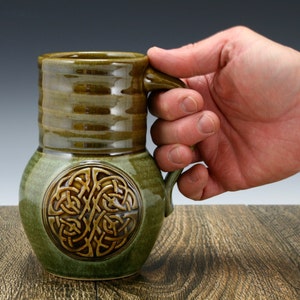 May include: A green ceramic mug with a brown Celtic knot design. The mug has a wide, rounded base and a handle.