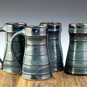 May include: Four handcrafted ceramic mugs with a dark teal glaze. Each mug features a cylindrical body with horizontal ridges and a large, curved handle. The mugs are arranged on a wooden surface, showcasing their unique, artisan design.