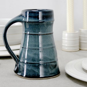 May include: A dark blue ceramic mug with a wide base and a rounded handle. The mug has a glazed finish and a slightly textured surface.
