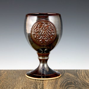 May include: A dark brown ceramic goblet with a Celtic knot design. The goblet has a wide base and a round bowl.