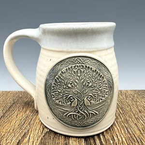 May include: Cream ceramic mug with a large handle and a grey Celtic tree of life design. The mug has a wide rim and a textured surface, suitable for hot drinks. The tree design is embossed on the mug's surface.