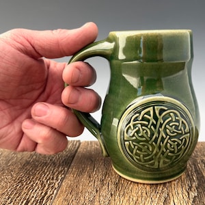 May include: A green ceramic mug featuring a Celtic knot design. The mug has a rounded body and a handle.