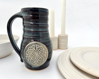 Puede incluir: Una taza de cerámica azul oscuro con un diseño de nudo celta en esmalte plateado. La taza tiene una asa ancha y un cuerpo redondeado.