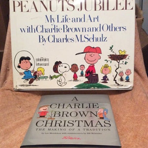 May include: Two vintage Peanuts books. The top book is titled "Peanuts Jubilee: My Life and Art with Charlie Brown and Others by Charles M. Schulz". The bottom book is titled "A Charlie Brown Christmas: The Making of a Tradition by Lee Mendelson with reminiscences by Bill Melendez".