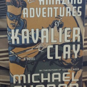 May include: Book cover for "The Amazing Adventures of Kavalier & Clay" by Michael Chabon. The cover features a comic book style illustration of a superhero in a blue and red costume.