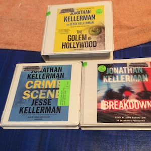 May include: Three white audio books with black text and colorful covers. The top book is titled "The Golem of Hollywood" by Jonathan Kellerman and Jesse Kellerman. The middle book is titled "Crime Scene" by Jonathan Kellerman and Jesse Kellerman. The bottom book is titled "Breakdown" by Jonathan Kellerman.