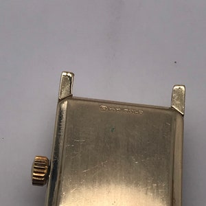 May include: Close-up of a vintage gold-toned watch. The watch has a rectangular face with a gold crown and lugs. The text "14K GOLD" is stamped on the watch face. The watch is likely from the mid-20th century.