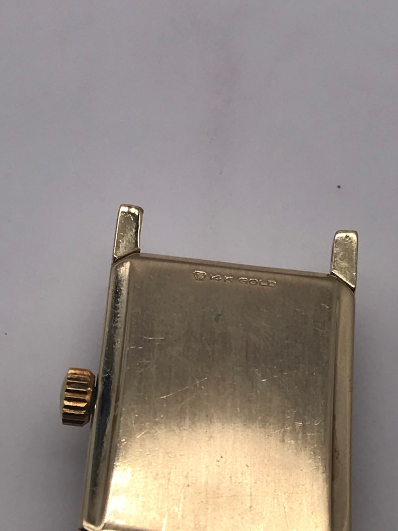 May include: Close-up of a vintage gold-toned watch. The watch has a rectangular face with a gold crown and lugs. The text "14K GOLD" is stamped on the watch face. The watch is likely from the mid-20th century.