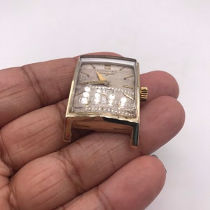 May include: A vintage gold-toned Longines wristwatch with a rectangular face. The watch has a light-colored dial with gold hands and hour markers. The watch is held in a hand against a white background.