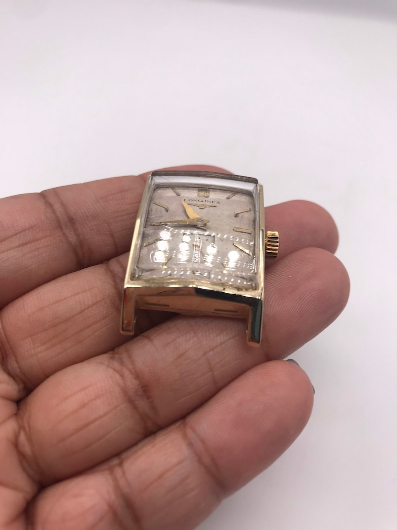 May include: A vintage gold-toned Longines wristwatch with a rectangular face. The watch has a light-colored dial with gold hands and hour markers. The watch is held in a hand against a white background.