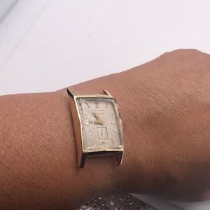 May include: A gold-toned rectangular wristwatch with a cream-colored face. The watch features a small subdial and the brand name "Longines" is visible. The watch is on a person's wrist.